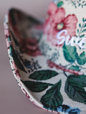 A macro close-up of a floral bucket-style hat brim, showing richly printed botanical fabric in cream, dusty rose, and deep green with a braided edge trim and partial embroidered script lettering.