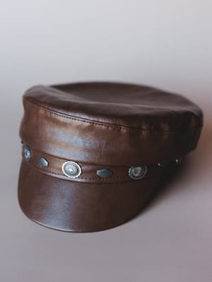 A rich brown faux leather captain's hat with a flat brim and a band of silver concho and diamond-shaped studs — the kind of hat that does half the outfit's work for you.