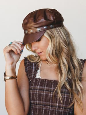 A close-up lifestyle shot of a model tilting the brim of a rich brown faux leather newsboy cap, styled with a smocked plaid top and layered gold jewelry — the whole look has that effortless 70s-leaning boho energy.