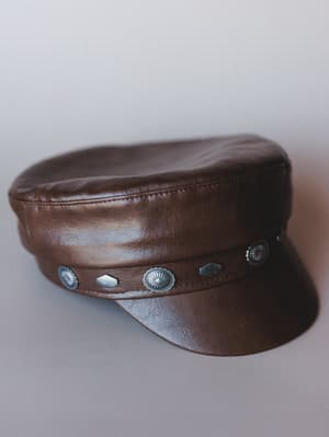A rich brown faux leather captain's hat with a flat brim and a band of alternating silver concho and geometric studs — the kind of hat that makes a simple outfit feel like a whole personality.