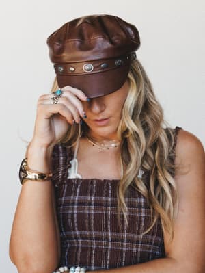 A close-up portrait of a model tipping the brim of a brown leather newsboy cap studded with silver conchos, paired with a smocked plaid top and layered boho jewelry — the whole look feels like a vintage record store meet a southwest road trip.