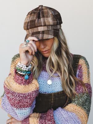 A layered, texture-rich look built around a brown plaid pageboy hat — the model tilts the brim low while a chunky multicolor stripe knit cardigan, light blue knit top, and stacked bead bracelets do the talking.