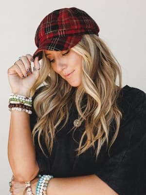A blonde model tips the brim of a red plaid pageboy hat with a soft, downward glance — the stacked bead bracelets, layered rings, and gold pendant necklace doing as much storytelling as the hat itself.