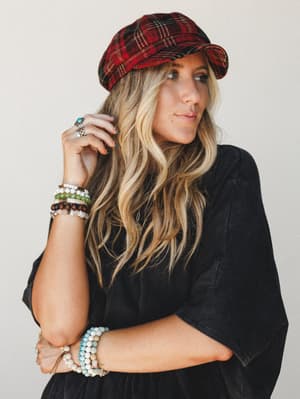 A relaxed, expressive shot of a woman in a washed black top and red plaid pageboy cap, her wrists stacked with beaded bracelets — the whole look reads like a fall Saturday with good intentions.