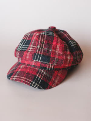 A red tartan newsboy cap laid flat on a soft grey surface — the kind of hat that makes a whole outfit feel intentional without trying too hard.