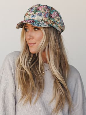 A soft, dreamy close-up of a model wearing a floral tapestry newsboy cap — the kind of hat that makes a cozy sweater look intentional and a coffee run feel like an event.