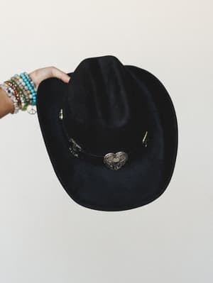 A black felt cowboy hat with a silver heart concho band is held up against a cream studio wall by a hand stacked with colorful beaded bracelets — effortlessly western with just enough personality.
