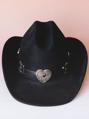 A black felt cowboy hat shot against a soft blush backdrop, centered on a silver engraved heart concho and leather band — western with just enough romance to make it feel personal.