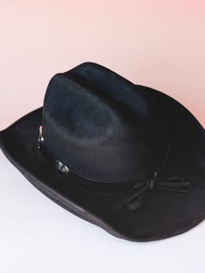 A black felt cowboy hat laid at a casual angle against a soft blush backdrop — the concho hardware and leather tie band give it that worn-in western charm that works just as hard at a concert as it does on a weekend errand.