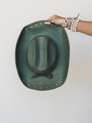 A distressed olive green cowboy hat held up from behind, showing the underside of the brim — studded edge detail and a leather tie at the crown, held by a hand stacked with beaded bracelets against a soft cream wall.