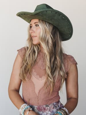 A dreamy mid-shot of a model in a olive suede wide-brim cowgirl hat with stud trim, layered over a dusty rose lace top and floral skirt — the kind of outfit that makes a country concert feel like a mood.