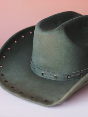 A close-up of a worn-in olive suede cowboy hat laid flat, with brass eyelet trim along the brim and a matching leather band — the kind of hat that already looks like it has a story.