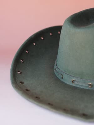 A close-up of a sage-olive felt cowgirl hat with brass grommet detailing along the brim and a matching hatband, shot against a soft dusty rose gradient background — the kind of hat that makes a whole outfit feel intentional.