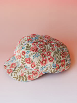 A floral tapestry-style baseball cap laid flat against a soft pink gradient background — the kind of hat that makes a simple outfit feel like it has a story.