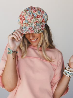 A smiling model tips the brim of a floral tapestry baseball cap with playful ease, paired with a blush pink sweatshirt and stacked beaded bracelets — the kind of effortless spring outfit that looks like it came together on the way out the door.