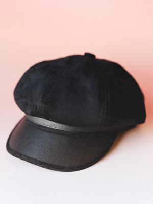 A black suede-textured newsboy cap with a faux leather brim and band detail, photographed against a soft blush pink backdrop — the kind of hat that makes a plain outfit feel intentional.