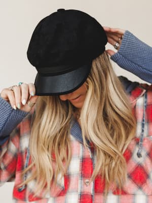 A model tilts the brim of a black suede baker boy cap with a faux leather brim, styled over wavy blonde hair alongside a red plaid shacket and a blue waffle-knit layer — the whole thing feels like a Saturday that starts with coffee and ends wherever.