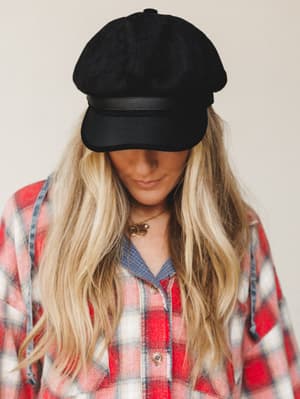 A close-up model shot spotlighting a black newsboy cap with a leather-trim brim, worn brim-down over the face with effortless cool, paired with a red plaid flannel and a delicate gold pendant necklace.