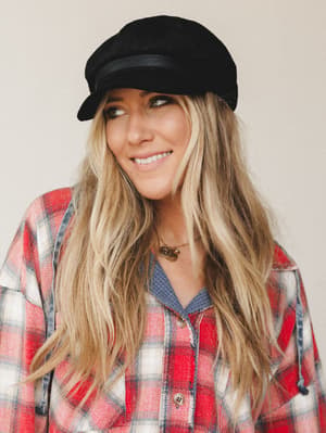 A smiling model in a black baker boy cap paired with a red plaid flannel and layered necklaces — the kind of effortless fall look that feels thrown together in the best possible way.