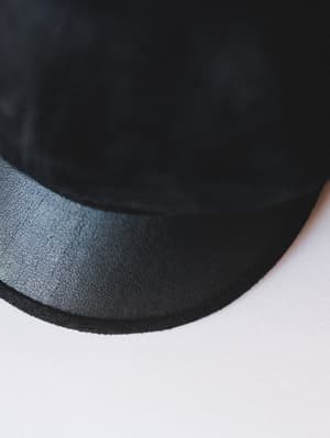 A close-up detail shot of a black hat with a structured leather-look brim, showing the contrast between the soft suede-like crown and the textured faux leather underside of the brim against a clean white surface.