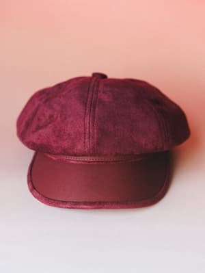 A rich burgundy newsboy cap shot against a soft blush backdrop — the suede crown and smooth faux-leather brim give it that vintage-meets-intentional energy that makes a whole outfit feel considered.