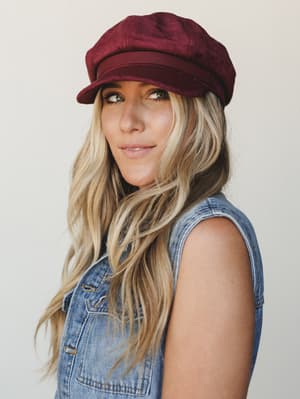 A close portrait of a model wearing a rich burgundy newsboy cap paired with a sleeveless denim vest — the kind of effortless fall outfit that looks like it happened on purpose.