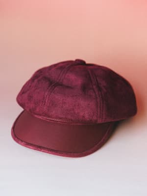 A rich burgundy newsboy-style cap shot against a soft pink gradient backdrop — the contrast of suede crown and faux leather brim gives it that intentional, lived-in vintage feel.