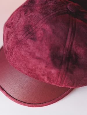 A close-up detail shot of a burgundy newsboy-style cap with a suede crown and contrasting faux leather brim, shot against a soft blush-white surface — the kind of hat that makes a whole outfit feel more intentional.