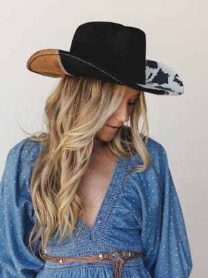 A blonde model looks softly downward wearing a statement black wide-brim cowboy hat with patchwork brim detailing — cow print, tan suede, and contrast stitching — paired with a blue polka dot smocked dress and a concho leather belt, all shot against
