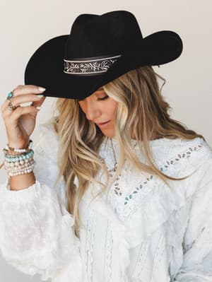 A model tilts the brim of a black felt cowboy hat trimmed with an embroidered floral band, paired with a white lace and eyelet top stacked with beaded bracelets — western charm meets boho softness.