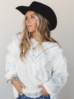 A white lace and ruffle top styled with a wide-brim black felt cowboy hat and stacked boho jewelry — romantic western dressing at its most lived-in.