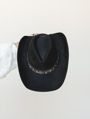 A black felt cowboy hat with a decorative floral-engraved band is held up against a soft cream wall, the white sleeve of the wearer peeking into frame — effortlessly western with a boho edge.