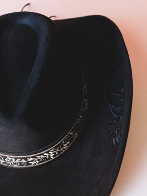A close-up of a black felt cowgirl hat with a floral embroidered hat band and tone-on-tone floral tooling on the brim, shot against a soft blush background — the kind of hat that makes any outfit feel intentional.