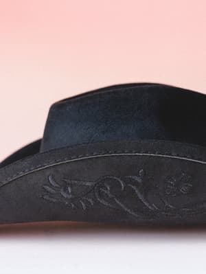 A close-up detail shot of a black suede cowboy hat resting against a soft blush backdrop, highlighting the tone-on-tone embroidered brim with a delicate bird and floral motif.
