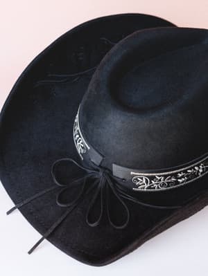 A black felt cowboy hat laid flat against a soft blush backdrop, its embroidered silver-and-black band and tied suede laces giving it that worn-in, Western-meets-boho personality.