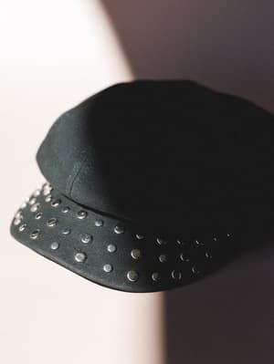 A black studded newsboy cap shot solo against a blush and shadow-split background — the silver hardware catches the light just enough to feel a little rock-and-roll without trying too hard.