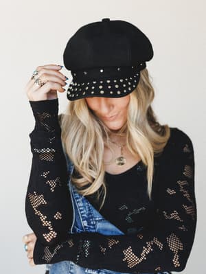 A blonde model tips the brim of a black studded newsboy hat with a layered-ring hand, wearing a black open-knit mesh sweater over denim overalls — the whole vibe is effortless rock-boho with a little attitude.