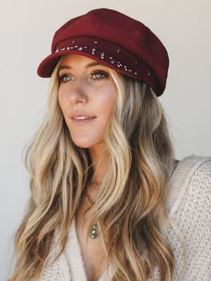 A close portrait of a model wearing a deep burgundy studded newsboy cap paired with a cream knit cardigan and layered gold necklaces — the kind of effortless fall look that feels intentional without trying too hard.