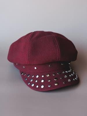 A deep burgundy newsboy cap with a suede-like texture and a brim covered in mixed silver studs — the kind of hat that makes a casual outfit feel intentional.