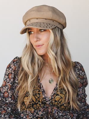A close-up portrait of a model in a khaki suede-look newsboy cap with silver stud detail along the brim, paired with a dark floral long-sleeve top and a layered gold coin necklace — the whole look has that effortless, lived-in boho energy.
