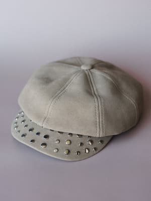 A khaki suede-look newsboy cap laid flat on a soft lavender surface, its structured panels and silver dome studs covering the brim giving it that lived-in-but-intentional energy.