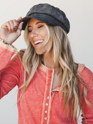 A smiling model tips a washed black newsboy cap with easy confidence, layered over a faded coral ribbed henley — the kind of effortless, thrown-together look that somehow always works.