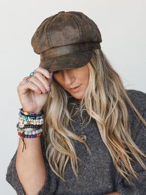 A close-up lifestyle shot of a model tipping the brim of a distressed brown faux-leather newsboy cap, her wrist stacked with colorful beaded bracelets and silver rings — the whole look feels like a rainy-day antique market in the best possible way.