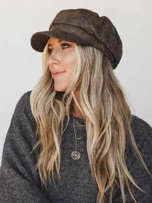 A close portrait of a woman in a distressed brown leather-look newsboy cap and charcoal sweater, layered gold necklaces catching the light — the whole thing has that effortless, I-just-threw-this-together energy that takes real style to pull off.