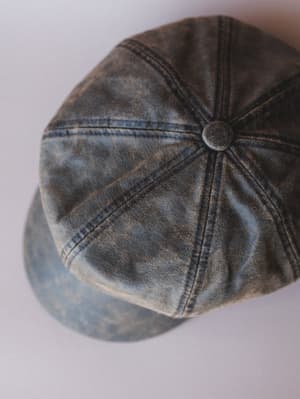 An overhead close-up of a washed newsboy cap in worn blue-brown tones, its panel seams and center button button catching soft studio light — the kind of hat that looks like it's already been somewhere interesting.