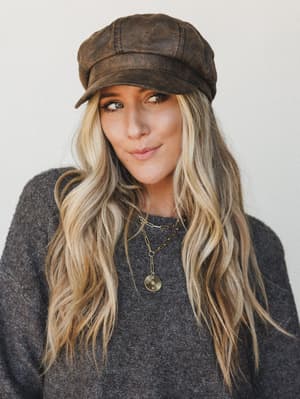 A close-up portrait of a model wearing a distressed brown faux-leather newsboy cap paired with a charcoal sweater and layered gold necklaces — the kind of effortless, lived-in look that makes a hat feel like a personality.