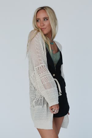 A woman with long blonde hair wears the Brenden Knit Netted Cardigan in ivory over a green top and black skirt, accessorized with layered necklaces and rings, standing against a plain white background.