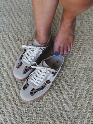 A close-up shot of leopard print sneakers on a jute rug — one shoe on, one off — with bright blue toenail polish adding an unexpected, personality-forward pop.