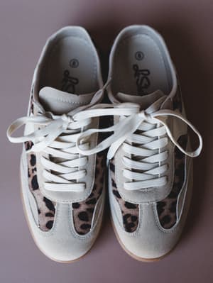 A pair of retro-style lace-up sneakers shot from above — cream suede paneling meets leopard print accents on a muted mauve background, giving off that 'I threw on something comfortable and somehow look great' energy.
