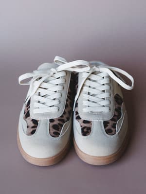 A pair of retro-style lace-up sneakers shot straight-on against a muted mauve background — cream suede uppers meet a leopard print panel in warm brown and black, grounded by a honey-toned gum sole.